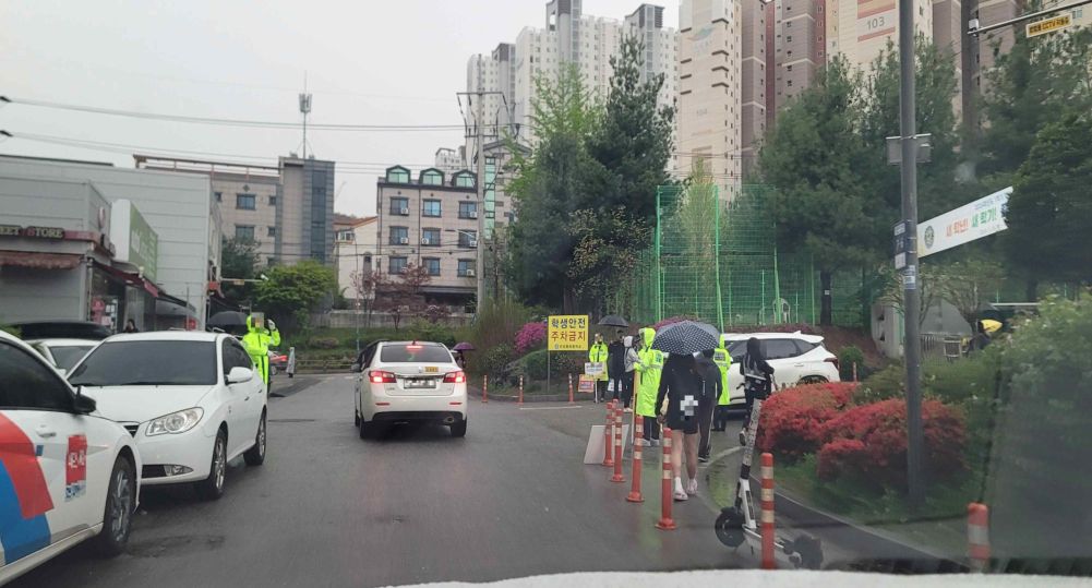 School commute on a rainy day · Back road without a centerline · Volunteer doing traffic safety work · Intersection without traffic signals · School main gate on the right
