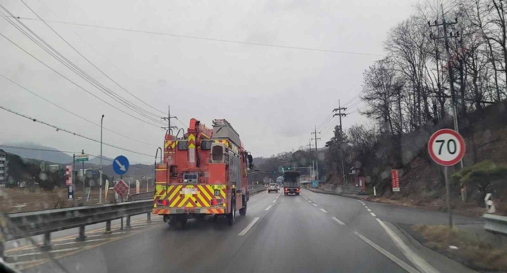 A – camera car, B – fire truck · Fire truck B photographed through the windshield of car A behind it · Raindrops falling, road surface wet · Crossroads intersection with traffic signals 300 meters ahead · Fire truck traveling in lane 1 with flashing lights and siren on