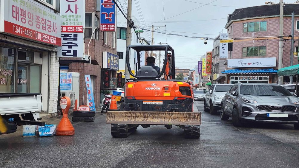 Piyodalar yo'lagi va qatnov qismi ajratilmagan turar-joy ichki ko'chasi · Yomg'irdan yo'l qoplamasi ho'l · Oldingi qurilish mashinasi sekin harakatlanmoqda · Yo'lning o'ng tomonida avtomobillar turibdi