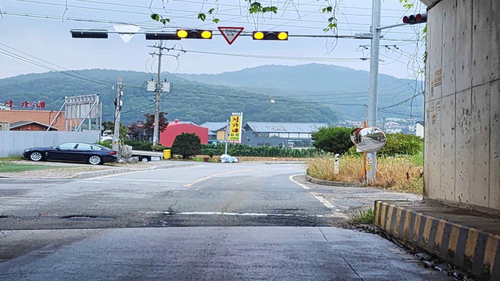 Vehicle traffic light flashing yellow · Safety sign reading "Slow" · Convex mirror on the right side of the road · Road surface is wet from rain · Potholes here and there on the road surface